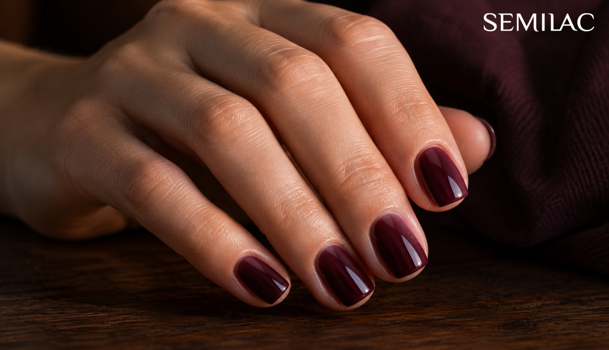 Hand with short squared natural nails wearing deep translucent blackberry gel polish, resting on dark wood beside plum knitwear